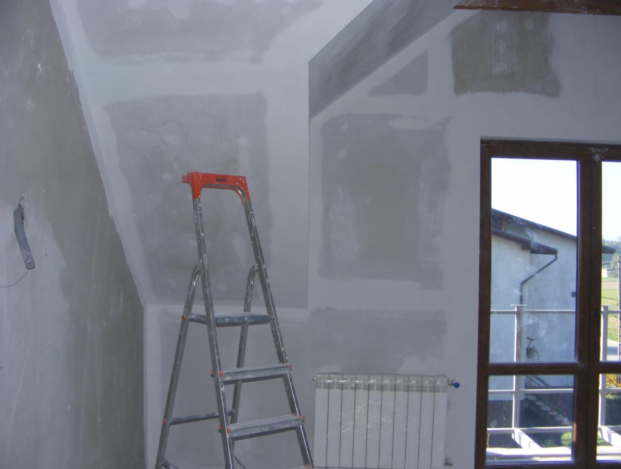 Poddasze w trakcie szpachlowania: ściany pokryte gładzią, drabina aluminiowa z pomarańczowymi elementami, okno z widokiem na budynek, grzejnik z folią ochronną.