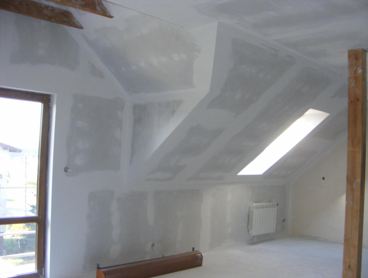 Poddasze ze skośnym sufitem w trakcie szpachlowania, widoczne płyty gipsowo-kartonowe i okno dachowe. Grzejnik na ścianie. W tle widok z okna.