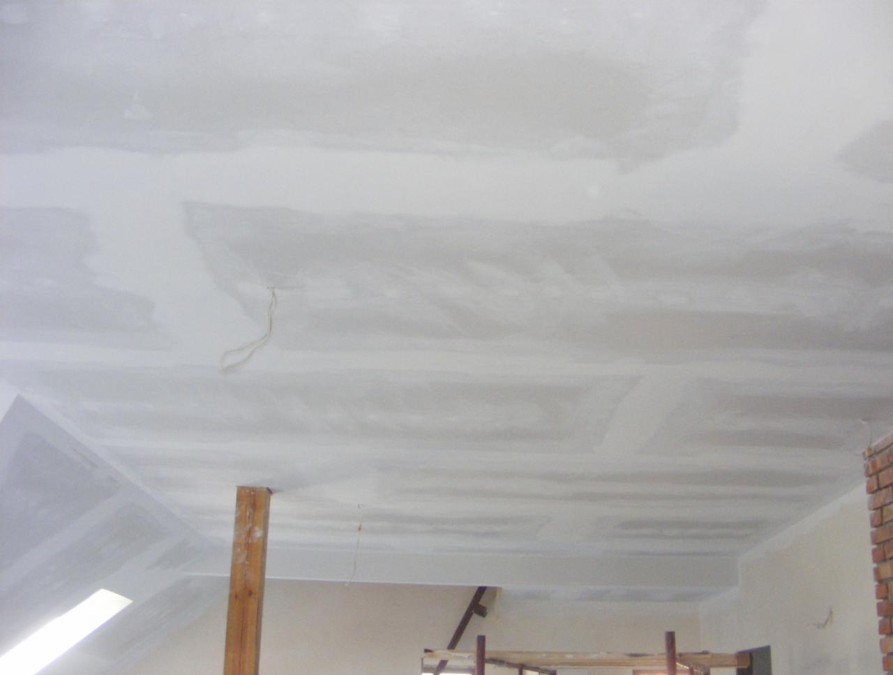 Nierównomiernie wyszpachlowany sufit poddasza z widocznymi fragmentami instalacji elektrycznej i drewnianą podporą, ukazujący etap prac wykończeniowych.