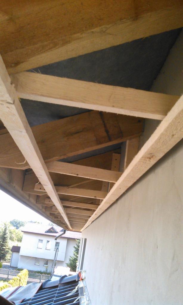 Odsłonięta konstrukcja drewniana okapu dachu, widok z dołu, z fragmentem elewacji budynku i dachu sąsiedniego budynku w tle.