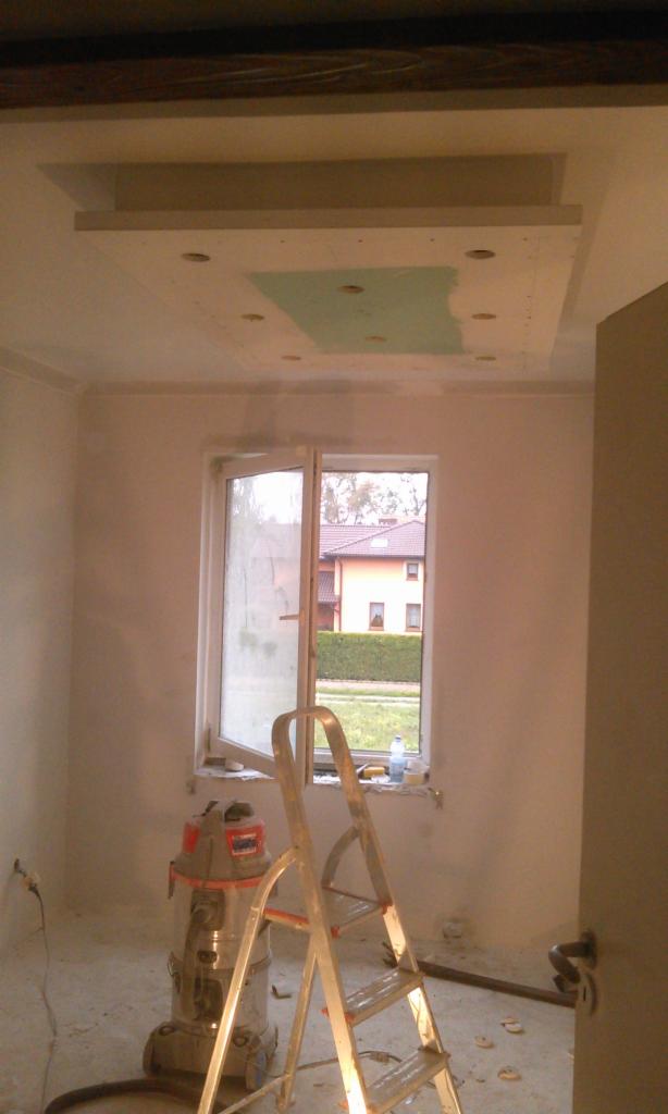 Pokój w trakcie remontu: sufit z podwieszaną konstrukcją i otwory na oświetlenie, drabina oraz odkurzacz przemysłowy przy oknie. Ściany przygotowane do malowania.