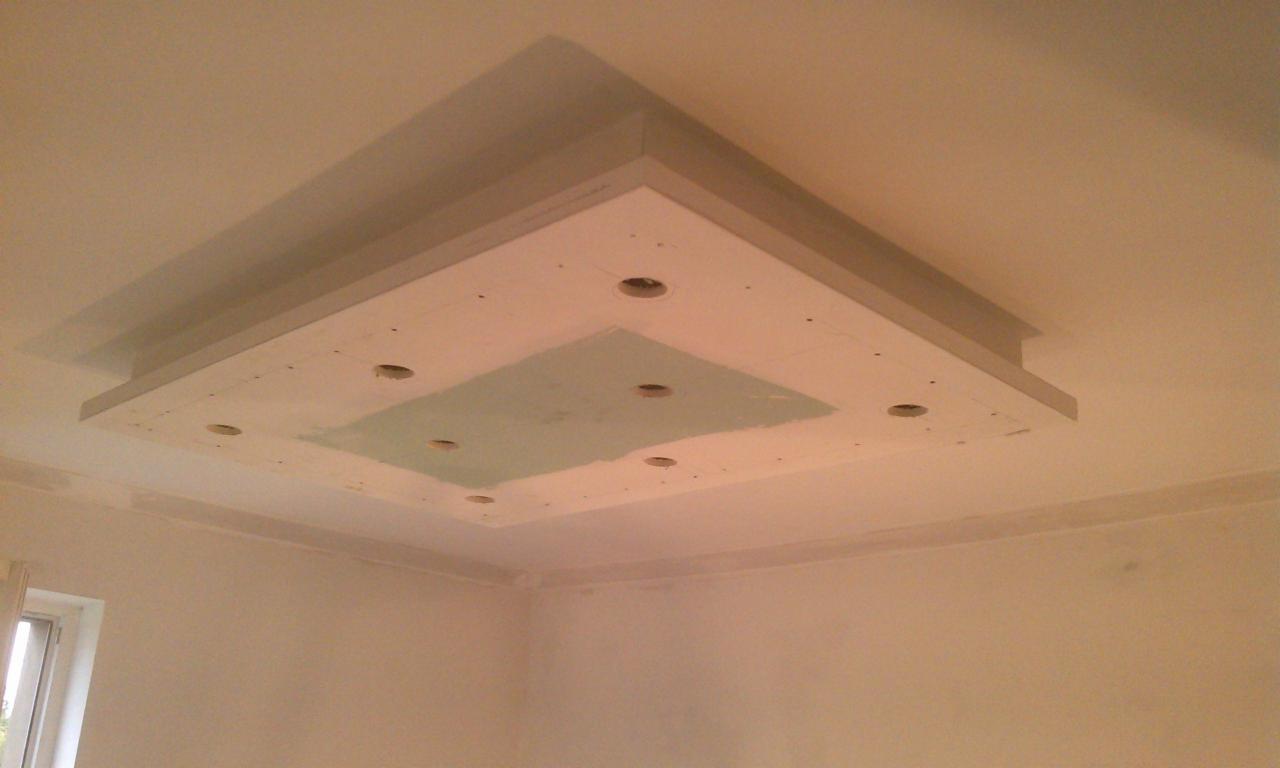 Sufit podwieszany z płyt gipsowych w trakcie montażu, widoczne otwory na oświetlenie i fragmenty szpachlowania, surowe ściany w tle.