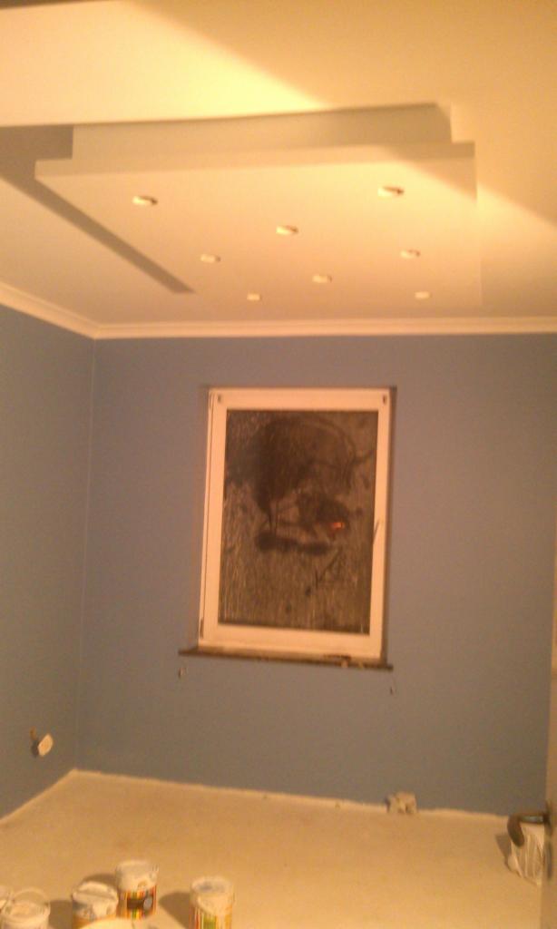 Pomalowane na niebiesko wnętrze pokoju z oknem, widoczne puszki z farbą i sufit z oświetleniem punktowym. Świeżo wykończone ściany.