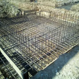 Siatka zbrojeniowa w wykopie fundamentowym, z widocznymi drewnianymi szalunkami i wystającymi pionowymi prętami, przygotowana do zalania betonem.