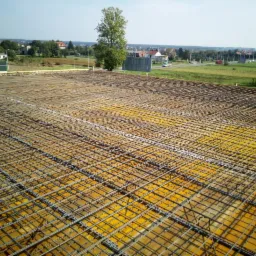 Siatka zbrojeniowa ułożona na żółtym szalunku na budowie z widokiem na oddalone zabudowania i zieleń.