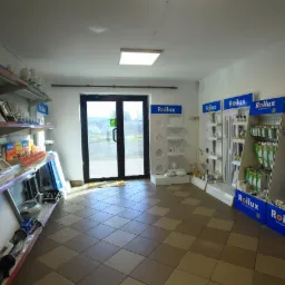 Wnętrze sklepu z oświetleniem Roilux: półki z lampami, żarówkami i panelami LED. Ekspozycja produktów oświetleniowych na tle przeszklonych drzwi wejściowych.