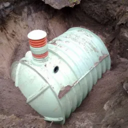 Zielony zbiornik retencyjny z pomarańczowym kominem, umieszczony w wykopie ziemnym. Widoczne ślady ziemi na zbiorniku i ścianach wykopu. Ujęcie z góry.