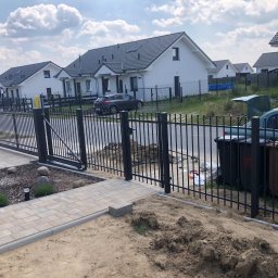 RobMaBud - Nowoczesne, czarne ogrodzenie panelowe z bramą przesuwną w trakcie montażu, widoczne przygotowane podłoże, kostka brukowa i elementy krajobrazu w tle.