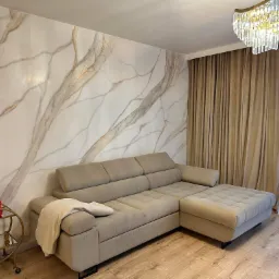 Salon z narożnikiem w odcieniach beżu, ściana z efektem marmuru i dekoracyjny żyrandol kryształowy.