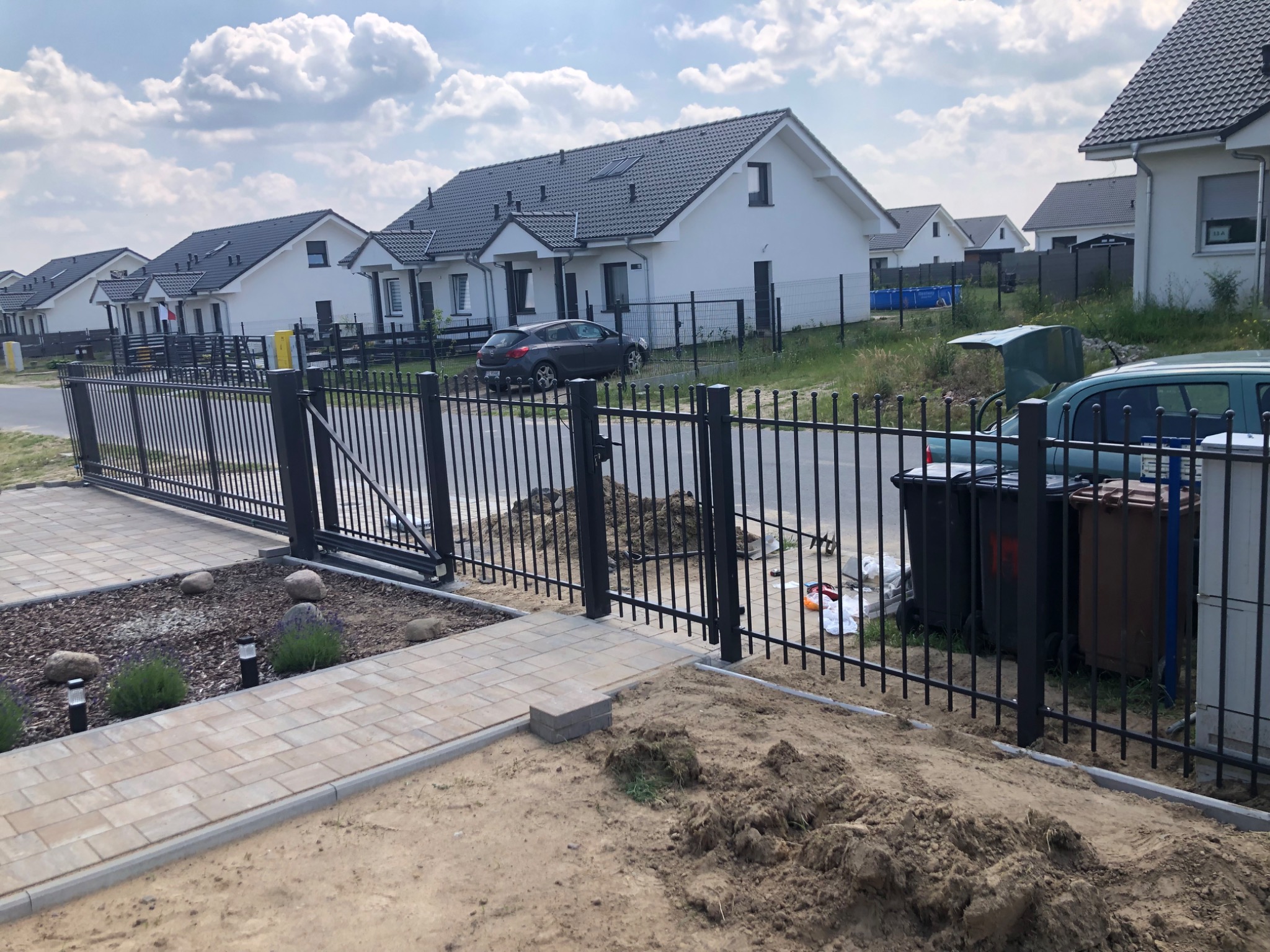 Nowoczesne, czarne ogrodzenie panelowe z bramą przesuwną w trakcie montażu, widoczne przygotowane podłoże, kostka brukowa i elementy krajobrazu w tle.