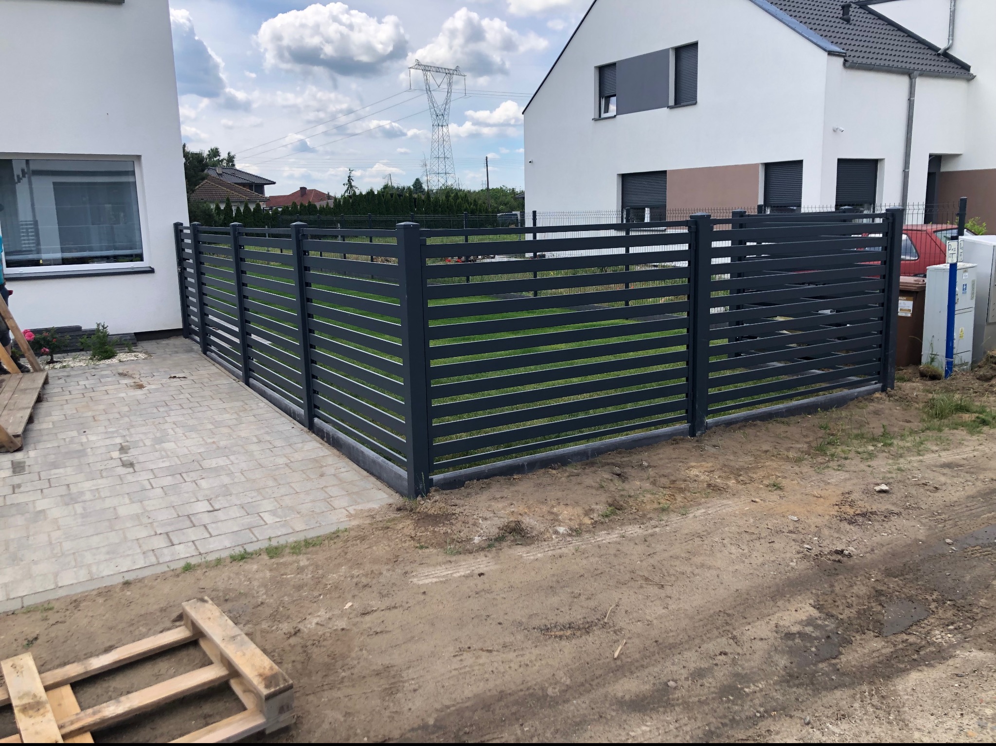 Nowoczesne, poziome ogrodzenie panelowe w kolorze grafitowym, oddzielające posesję z kostką brukową od niezagospodarowanego terenu z widoczną paletą drewnianą i słupem wysokiego napięcia w tle.