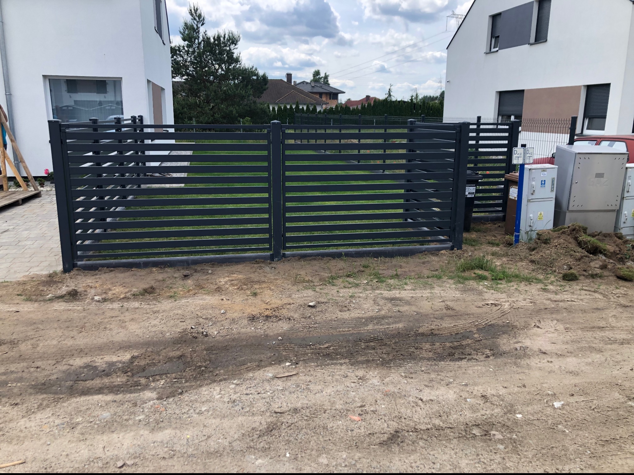 Nowoczesne, poziome ogrodzenie panelowe w kolorze grafitowym, oddzielające posesję z dwoma białymi budynkami mieszkalnymi, widoczne ślady prac ziemnych.