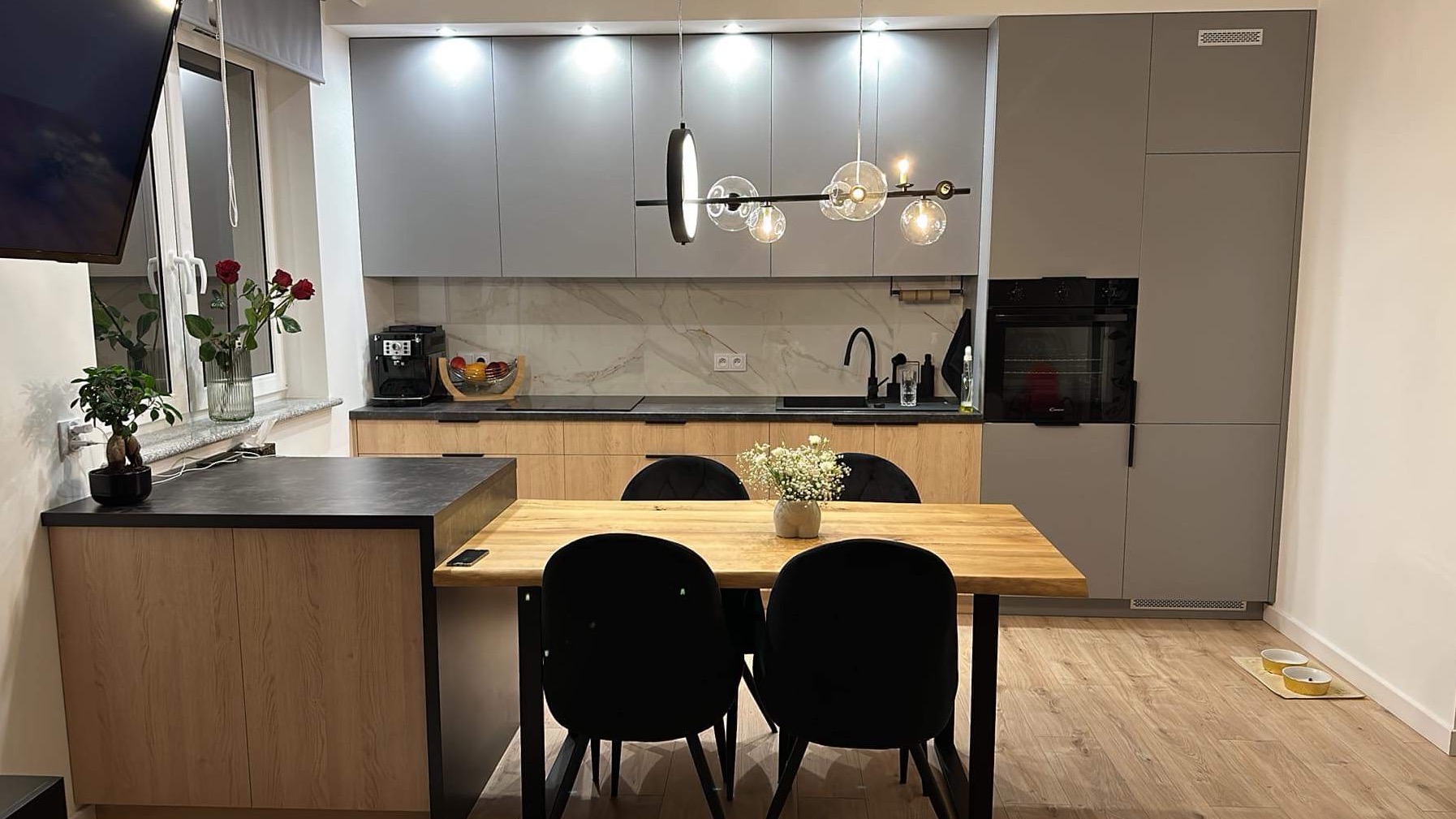 Nowoczesna kuchnia z szarymi szafkami, drewnianym blatem, marmurowym backsplashiem i designerską lampą wiszącą nad drewnianym stołem z czarnymi krzesłami.