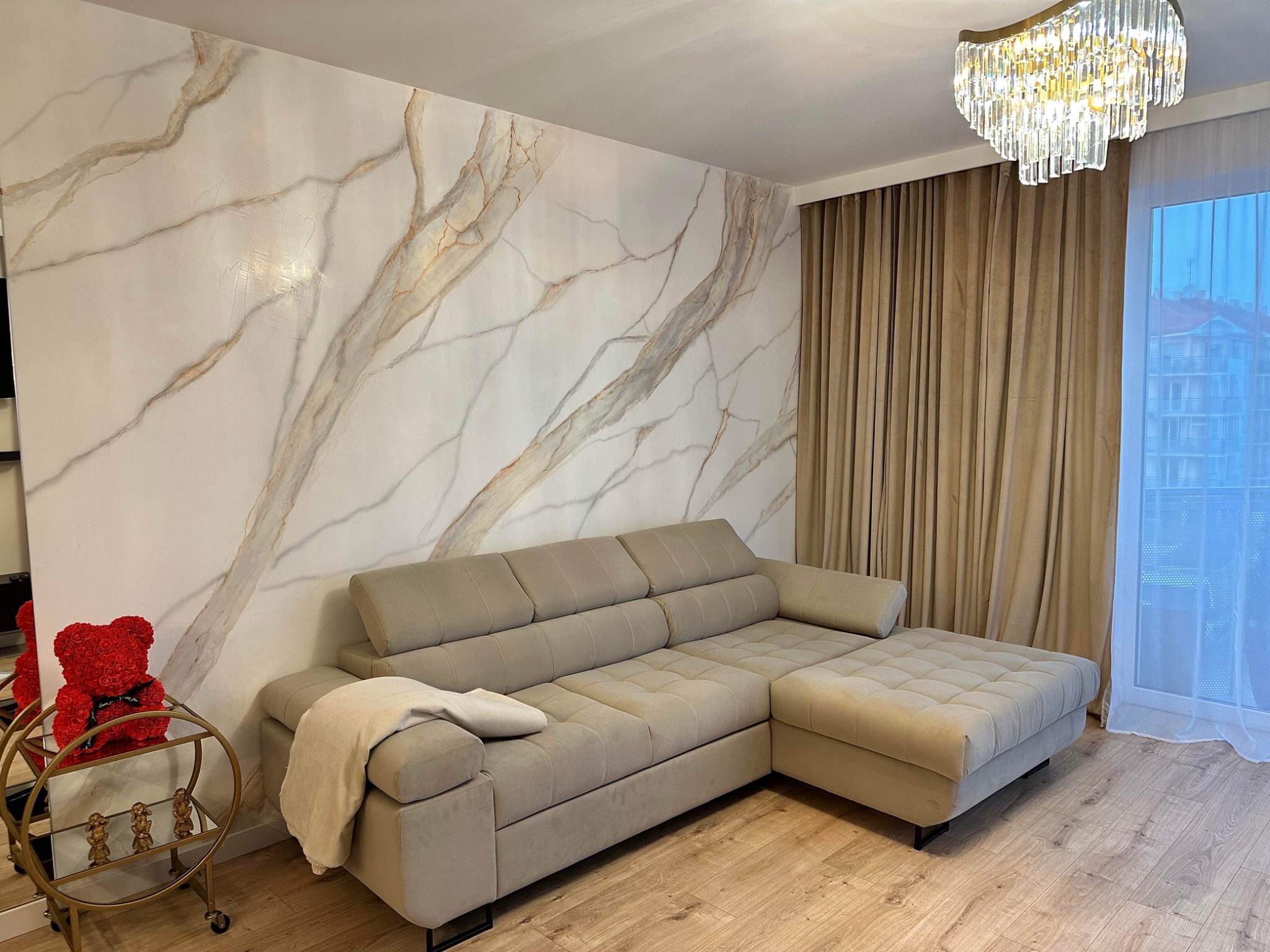 Salon z narożnikiem w odcieniach beżu, ściana z efektem marmuru i dekoracyjny żyrandol kryształowy.