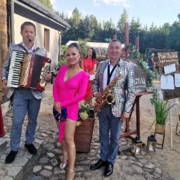 Zespół muzyczny w błyszczących marynarkach pozuje z akordeonem i saksofonem obok planu stołów na zewnątrz.