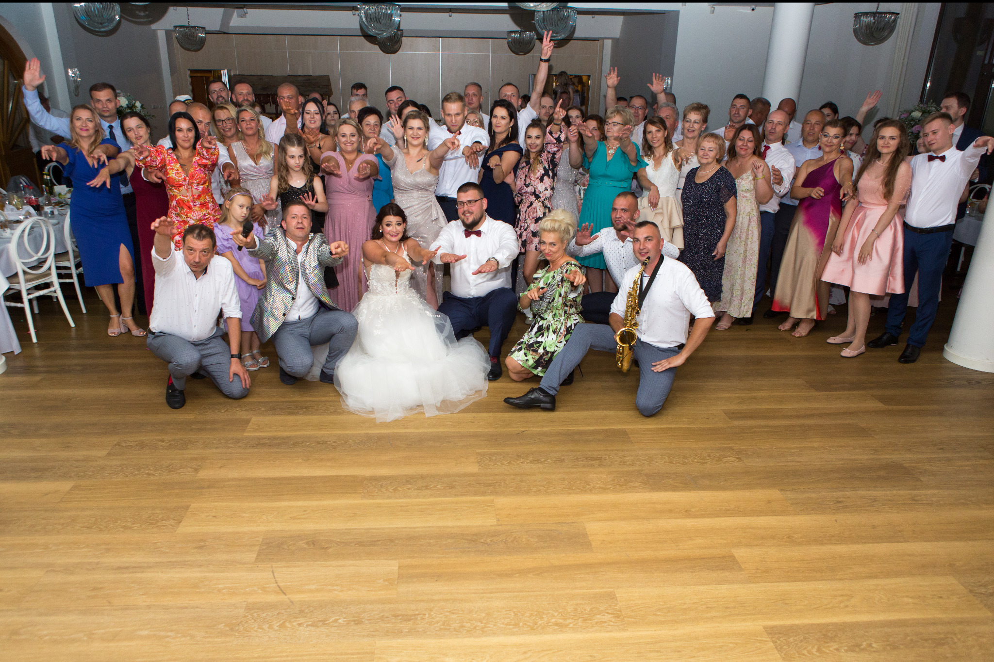 Zdjęcie grupowe gości weselnych z parą młodą w Wasilkowie, z saksofonistą klęczącym na pierwszym planie i parkietem z paneli podłogowych.