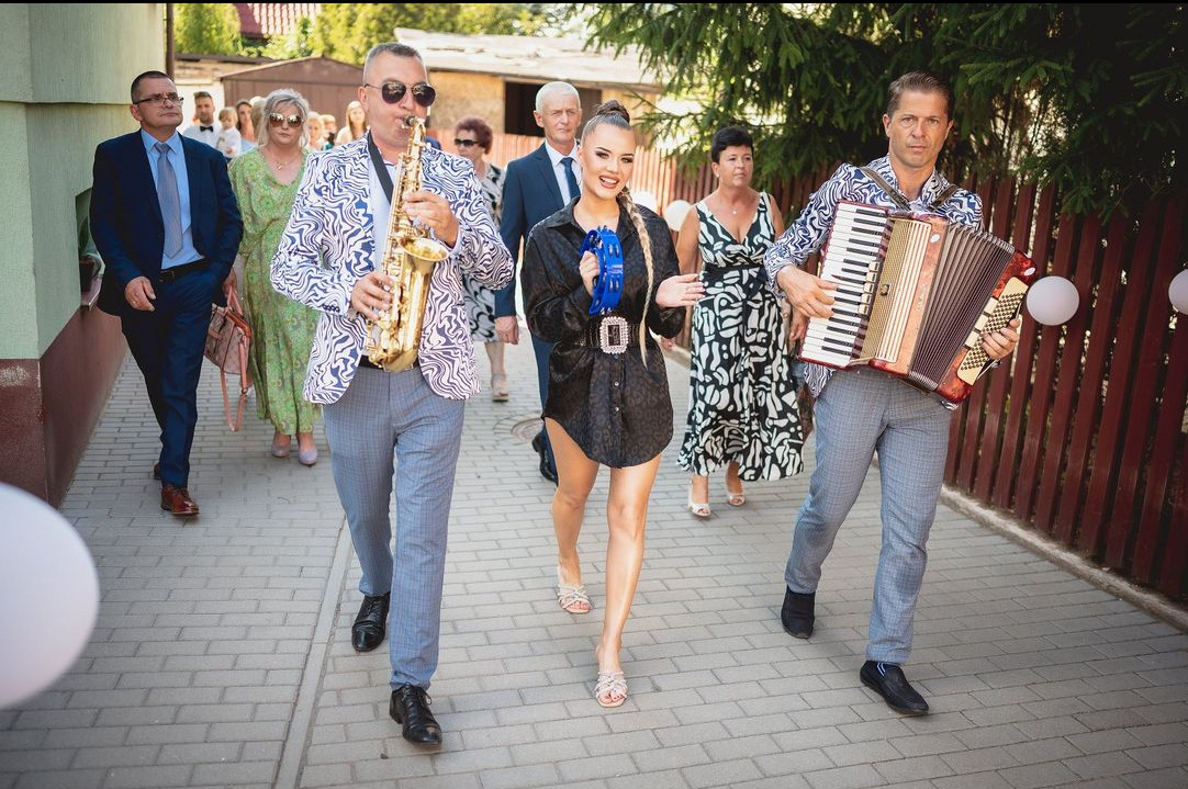Zespół muzyczny w trakcie przemarszu: saksofonista w wzorzystej marynarce, akordeonista i wokalistka z tamburynem prowadzą orszak weselny.
