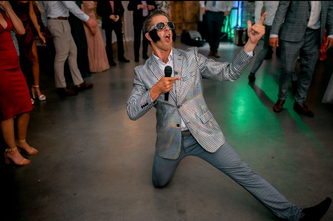 Mężczyzna w srebrnej, błyszczącej marynarce, okularach przeciwsłonecznych i sztucznych bokobrodach, klęczy i śpiewa do mikrofonu na tle bawiących się osób.