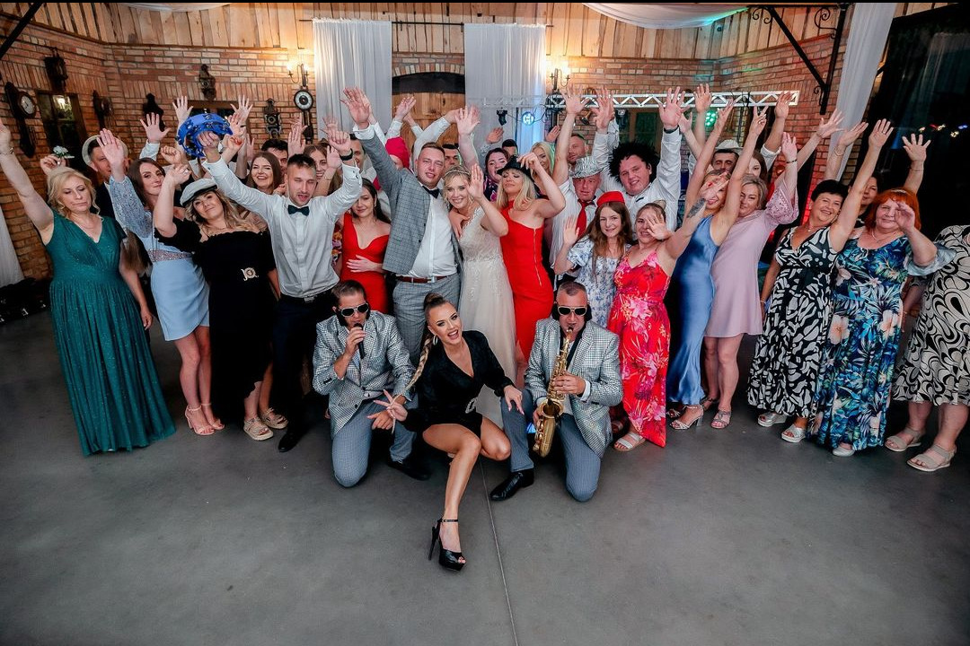 Duża grupa osób, w tym panna młoda w białej sukni, pozuje do zdjęcia z dwoma muzykami w błyszczących marynarkach, jeden z mikrofonem, drugi z saksofonem, na tle dekoracji sali weselnej...