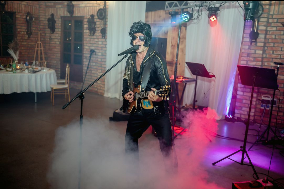 Mężczyzna w stroju Elvisa Presleya śpiewa i gra na gitarze na scenie z mikrofonem i efektami dymnymi, w tle dekoracje z cegły i zasłony.