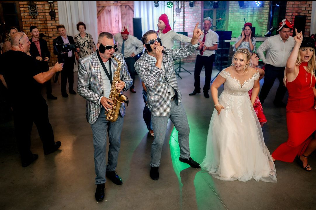 Duet muzyczny w kraciastych garniturach i okularach przeciwsłonecznych gra i śpiewa na weselu; saksofonista i wokalista, w tle tańczący goście w różnorodnych nakryciach głowy.