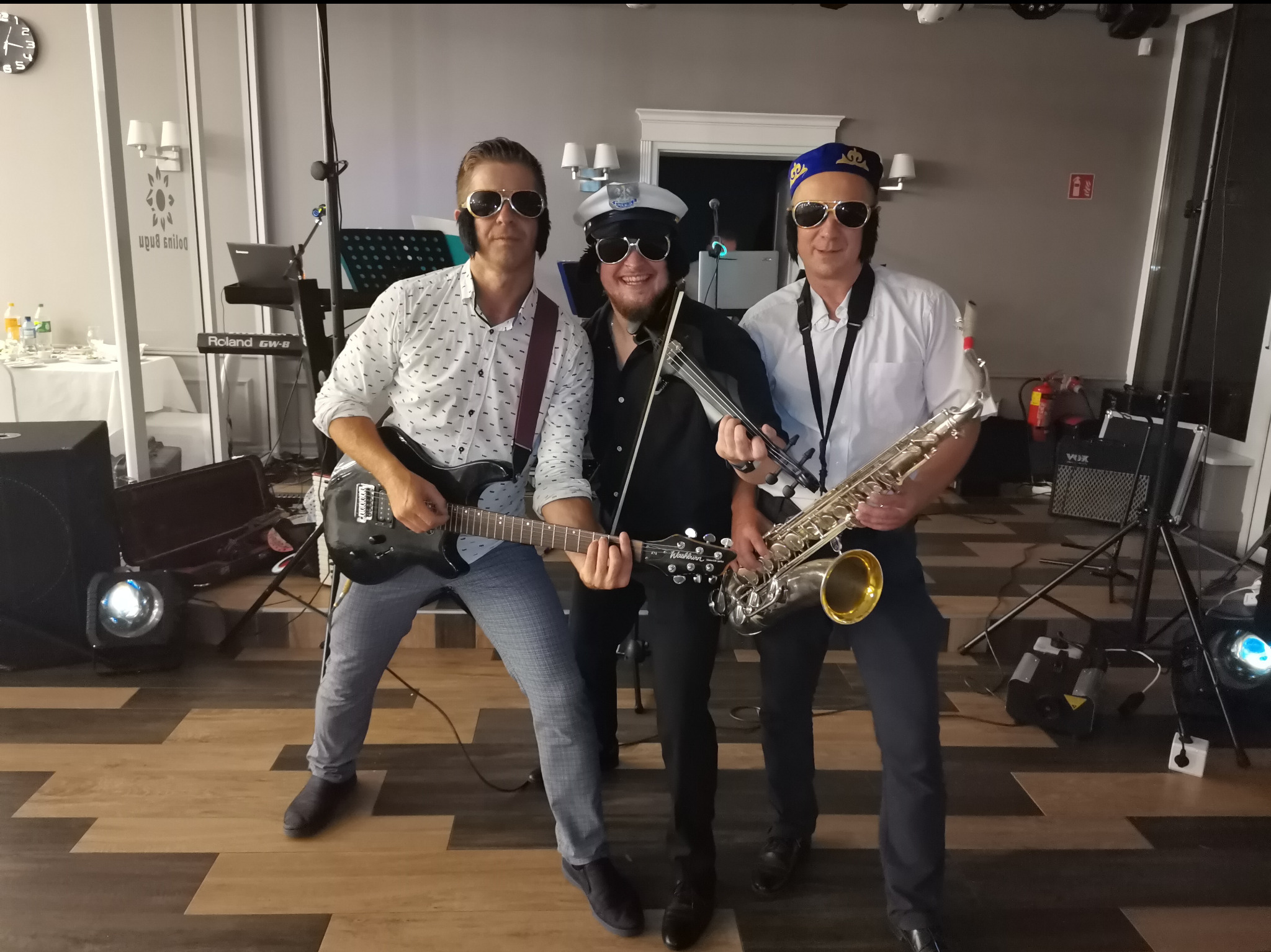 Trzech muzyków w okularach przeciwsłonecznych, jeden z gitarą elektryczną, drugi ze skrzypcami, a trzeci z saksofonem, w tle sprzęt nagłośnieniowy i dekoracje sali.