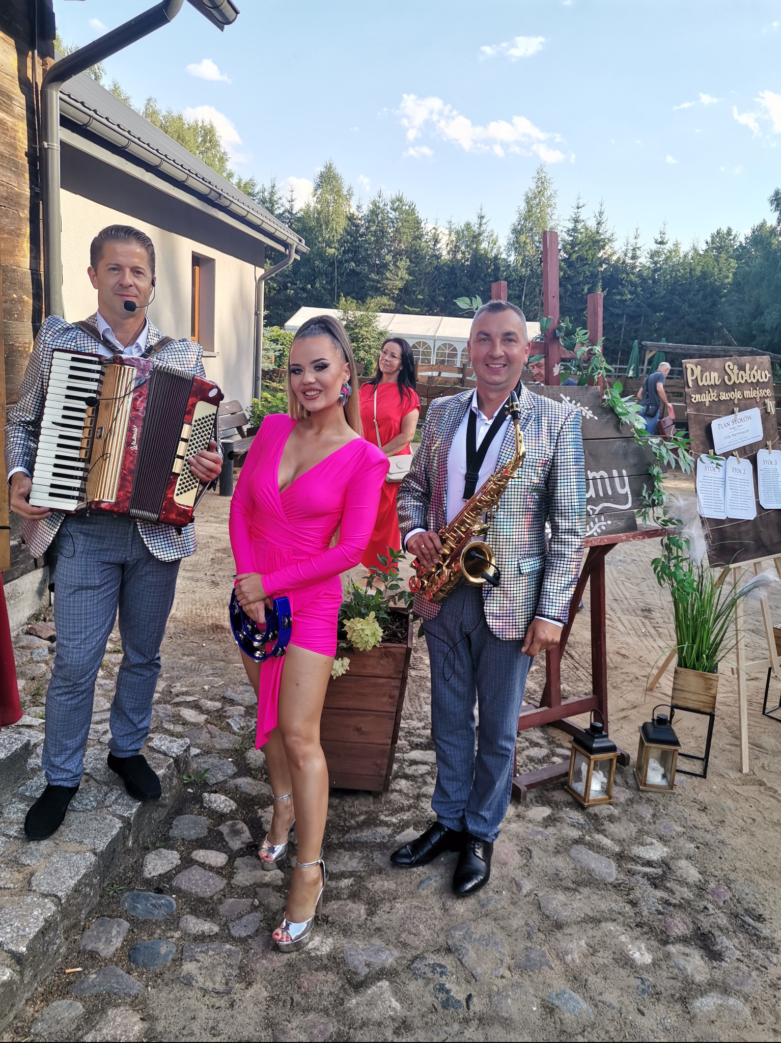 Zespół muzyczny w błyszczących marynarkach pozuje z akordeonem i saksofonem obok planu stołów na zewnątrz.