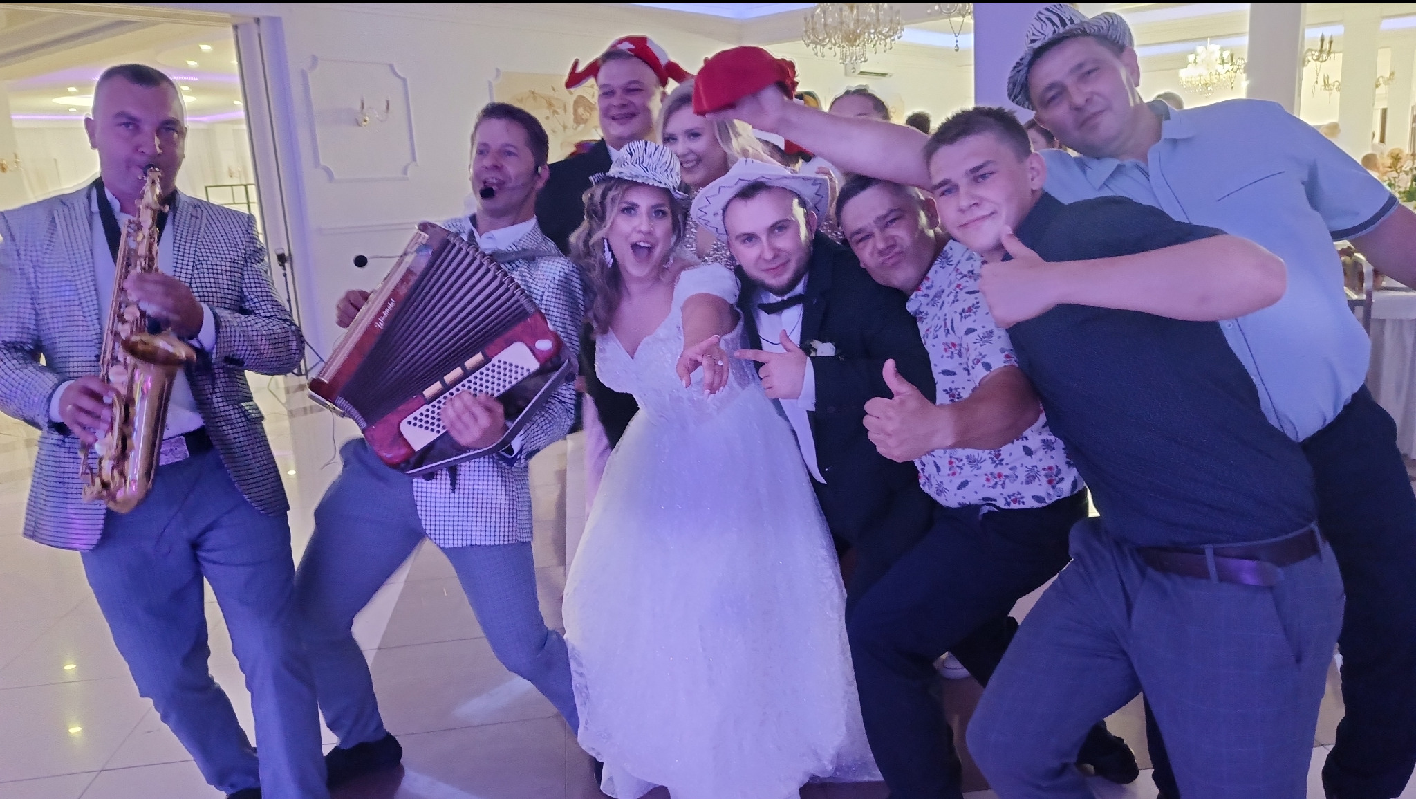 Zespół muzyczny w trakcie występu z nowożeńcami i gośćmi weselnymi, saksofonista i akordeonista w akcji, uśmiechnięta panna młoda w białej sukni.