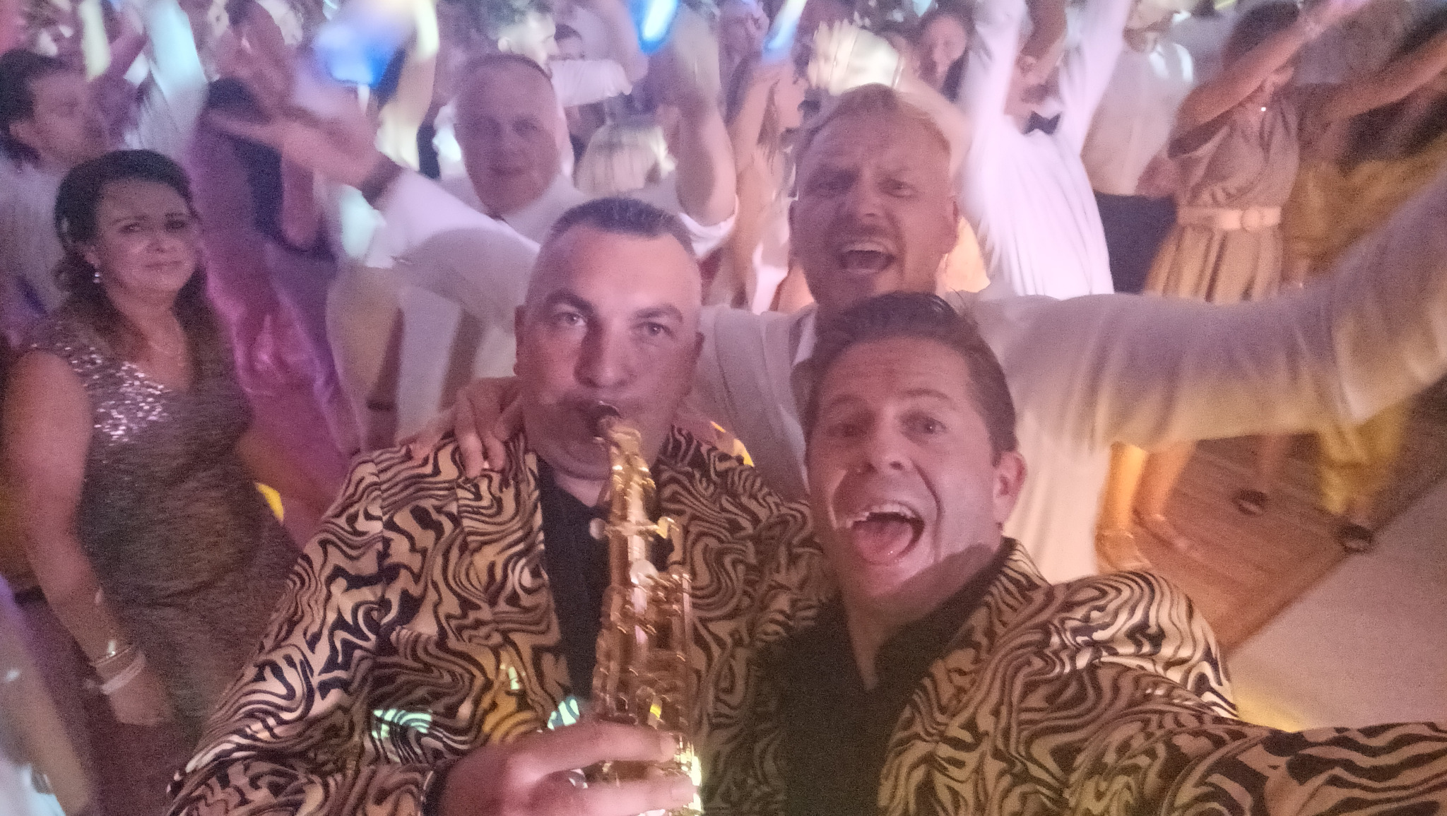 Saksofonista w marynarce w zebrę gra podczas imprezy, w tle rozbawieni goście w białych koszulach i sukienkach.