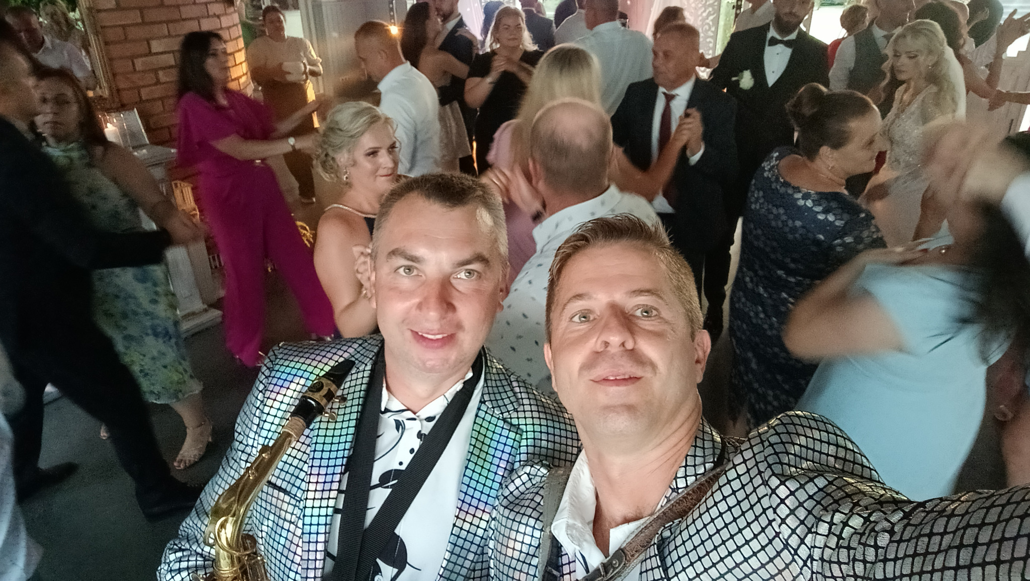 Selfie dwóch muzyków z saksofonem w błyszczących marynarkach na parkiecie podczas imprezy, w tle tańczący goście.