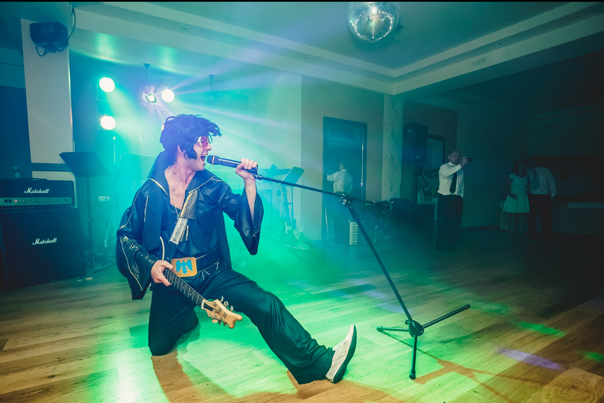 Imitator Elvisa Presleya w czarnym kostiumie, klęczący na scenie z gitarą i mikrofonem podczas występu na imprezie, z nagłośnieniem Marshall i dyskotekową kulą w tle.