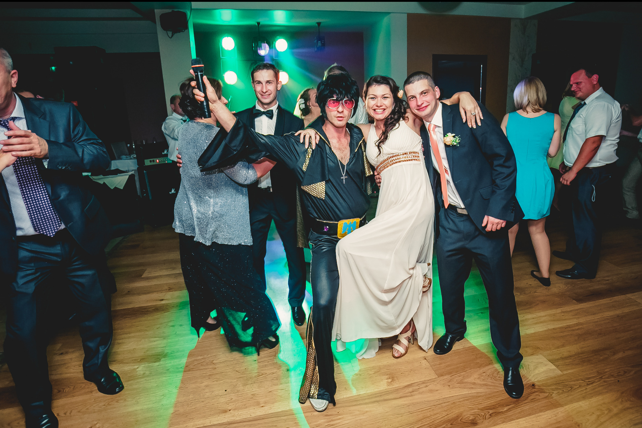 Impreza okolicznościowa: Osoba przebrana za Elvisa Presleya w czarnym kostiumie z mikrofonem pozuje do zdjęcia z gośćmi weselnymi na parkiecie.