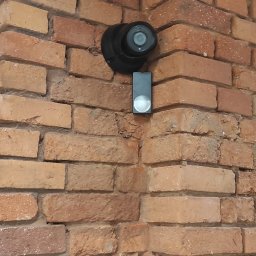 DP Technika - Czarna kamera monitoringu kopułkowa oraz czarny wideodomofon z okrągłym przyciskiem zamontowane na narożnej ścianie z czerwonej cegły.