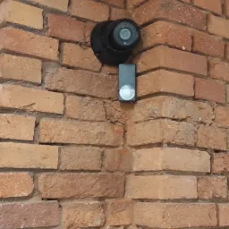 Czarna kamera monitoringu kopułkowa oraz czarny wideodomofon z okrągłym przyciskiem zamontowane na narożnej ścianie z czerwonej cegły.