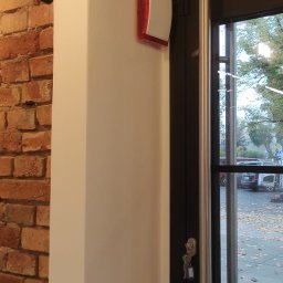 DP Technika - Wewnętrzna syrena alarmowa z czerwoną lampką na ścianie, obok drzwi wejściowych z kluczami w zamku, widok na zewnątrz z opadłymi liśćmi.