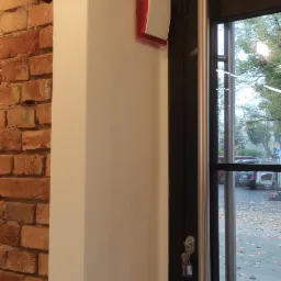 Wewnętrzna syrena alarmowa z czerwoną lampką na ścianie, obok drzwi wejściowych z kluczami w zamku, widok na zewnątrz z opadłymi liśćmi.