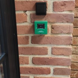 DP Technika - Zielony przycisk awaryjnego otwierania drzwi z napisem 'Naciśnij mocno do zadziałania' i czarny włącznik światła zamontowane na ceglanej ścianie w pobliżu ramy okiennej.