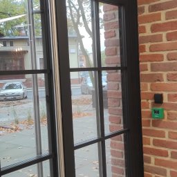 DP Technika - Czarne, przeszklone drzwi wejściowe z automatycznym systemem otwierania, zamontowane w budynku z cegły, widoczne zielone i czarne przyciski na ścianie obok drzwi.