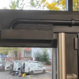 Zamknięcie automatyczne szarej, przeszklonej ramy drzwi z widocznymi czujnikami magnetycznymi i mechanizmem sprężynowym, na zewnątrz zaparkowany biały samochód serwisowy.