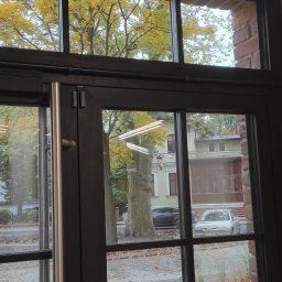 DP Technika - Widok z wnętrza budynku przez przeszklone drzwi z czarną ramą na ulicę z zaparkowanymi samochodami i kilkoma białymi furgonetkami z otwartymi drzwiami, ceglana ściana po prawej stronie.