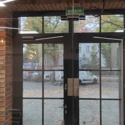 DP Technika - Szklane drzwi wejściowe z czarną ramą i metalowymi uchwytami, widok na ulicę z opadłymi liśćmi, nad drzwiami znak 'Wyjście ewakuacyjne' oraz klimatyzator Bosch.