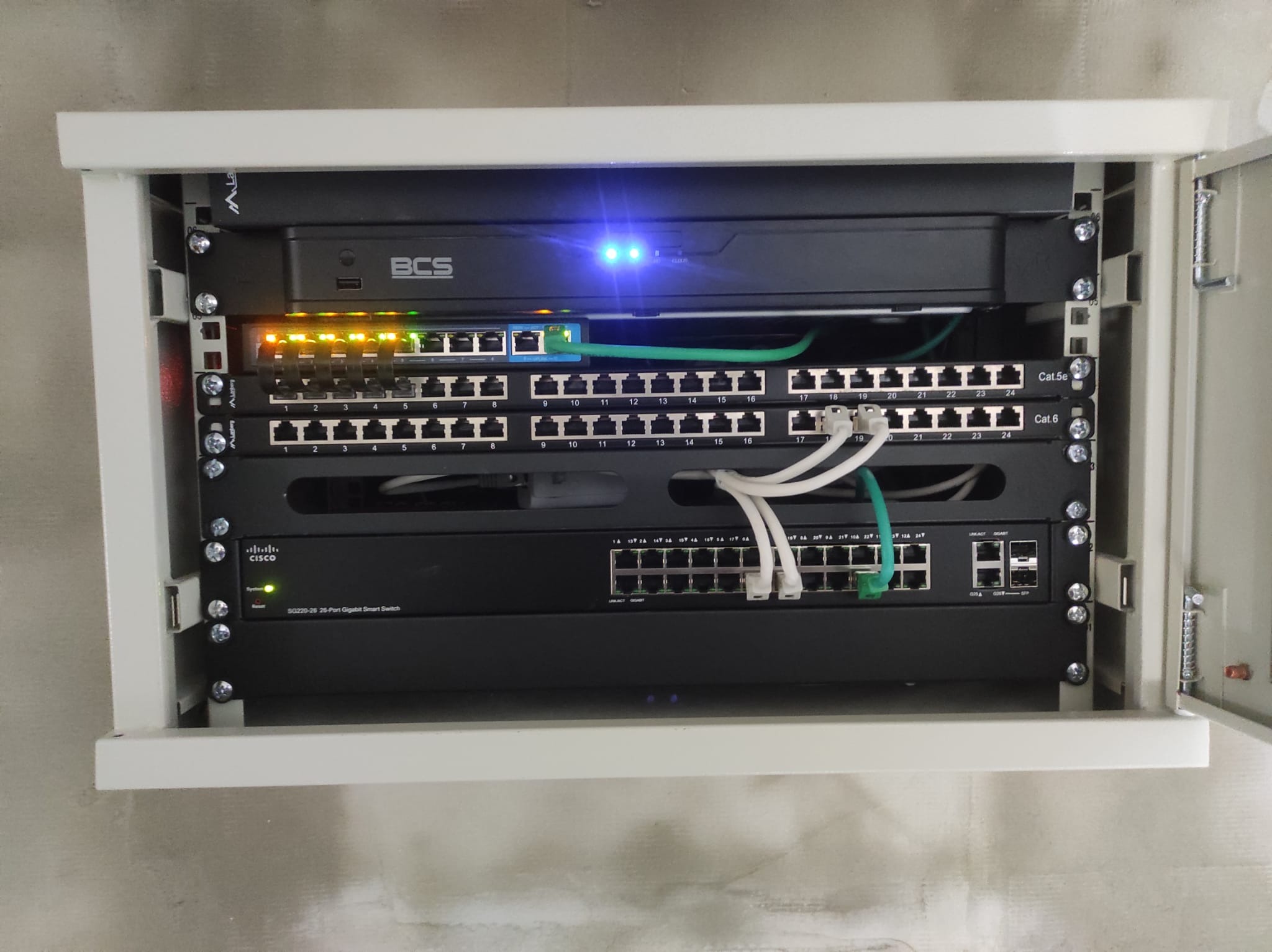 Szafa rack z zamontowanym rejestratorem BCS, przełącznikami sieciowymi Cisco i patch panelem Cat.5e oraz Cat.6, okablowanie strukturalne w kolorach białym i zielonym, diody sygnalizacyjne wskazują...