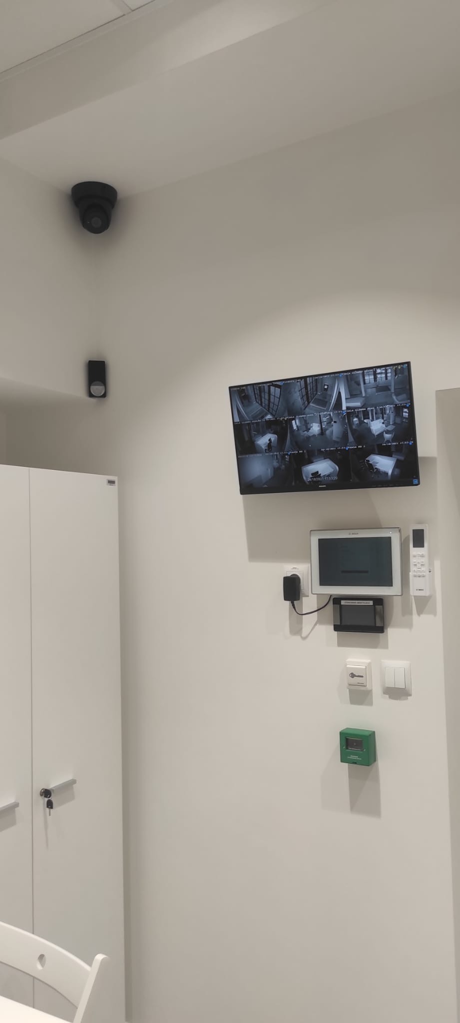 Wnętrze z zainstalowanym systemem monitoringu: kamera kopułkowa na suficie, czujnik ruchu, monitor z podglądem z kamer, panel sterowania i zielony przycisk alarmowy.