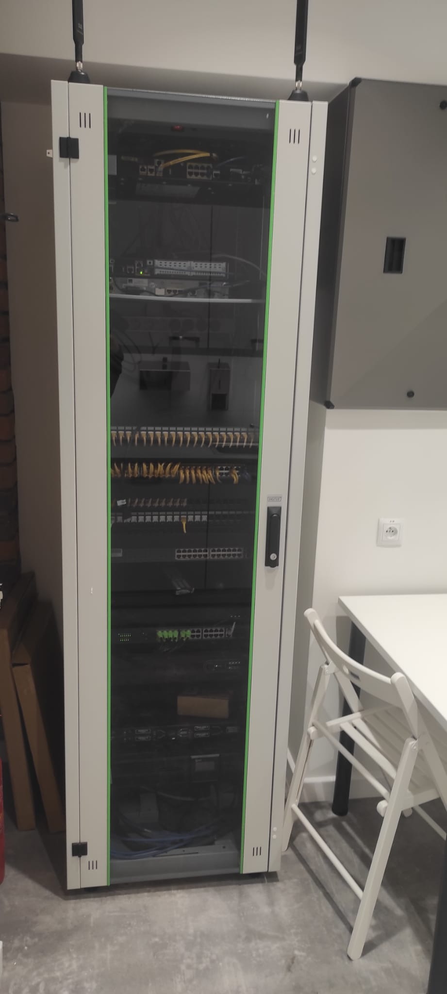 Zamknięta szafa rack z zainstalowanymi urządzeniami sieciowymi, widoczne patch panele, switche i okablowanie, obok biały stół i krzesło.