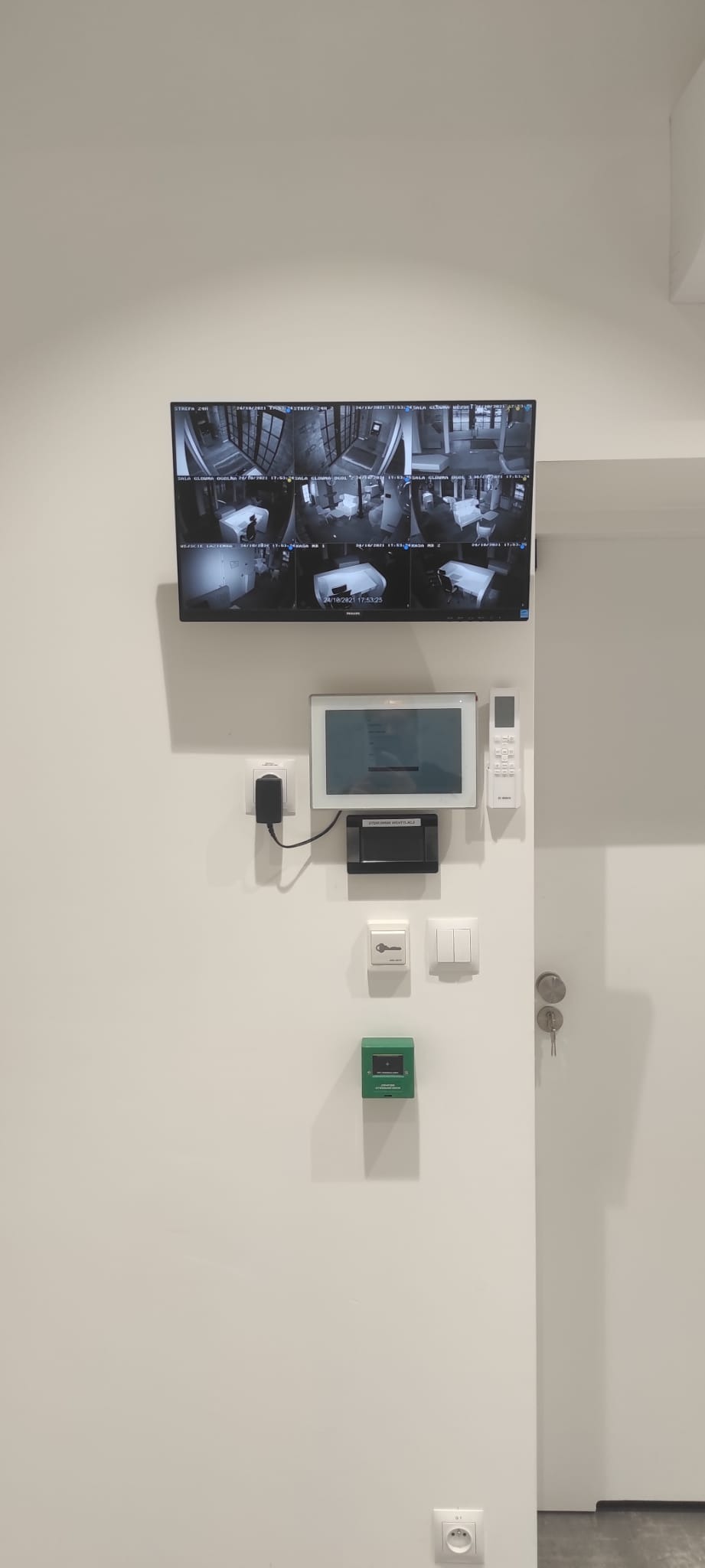 Ekran monitoringu z widokiem z sześciu kamer, panel sterowania, przycisk alarmowy i klucze na białej ścianie w pomieszczeniu.