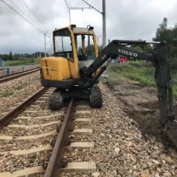 Żółta koparka Volvo na torach kolejowych, obok wykopana ziemia, pochmurne niebo w tle.