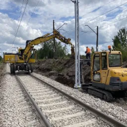 Żółta koparka kołowa na torach kolejowych wykonuje prace ziemne przy nowym słupie trakcyjnym, w tle widoczna mniejsza koparka i pracownik w pomarańczowej kamizelce.