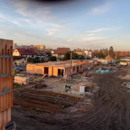 Budowa domu jednorodzinnego w trakcie wznoszenia ścian z czerwonej cegły, widok z góry na plac budowy z widocznymi elementami konstrukcyjnymi i materiałami budowlanymi, w tle zabudowa mieszkalna...