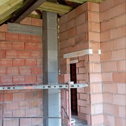 P.H.U Emilka Emilia Wołczyńska - Wnętrze w trakcie budowy: ściany z czerwonej cegły, szara kolumna, drewniany strop, widoczne rusztowanie i pusty otwór drzwiowy.