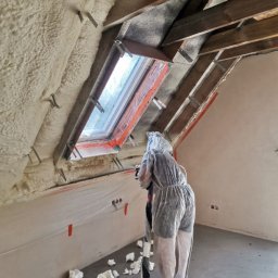 P.H.U Emilka Emilia Wołczyńska - Pracownik w kombinezonie ochronnym natryskuje pianę izolacyjną na poddaszu, widoczne krokwie, okno dachowe zabezpieczone folią, fragment ściany i resztki piany na podłodze.